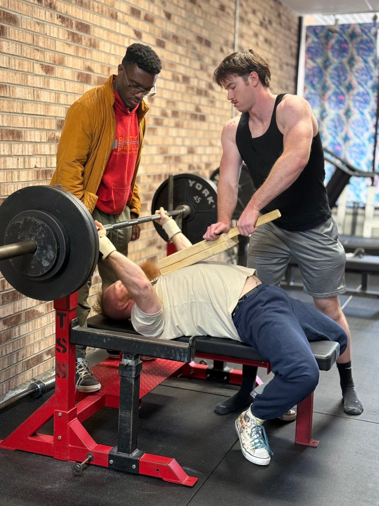 A man sets up for a bench press exercise while his coach and another gym goer assist.
